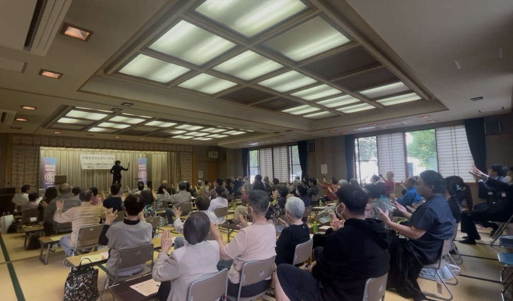 広い会場で、手を挙げて参加する大勢の聴講者を前に講演する藤田大知